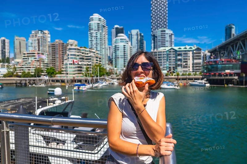 Young woman enjoying a delicious donut with the stunning cityscape of vancouver, british columbia, as her backdrop, showcasing a perfect blend of urban exploration and sweet indulgence