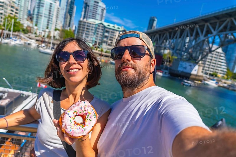Happy tourists taking a selfie while holding a donut with sprinkles in front of the cityscape and burrard bridge in vancouver harbor, british columbia, canada