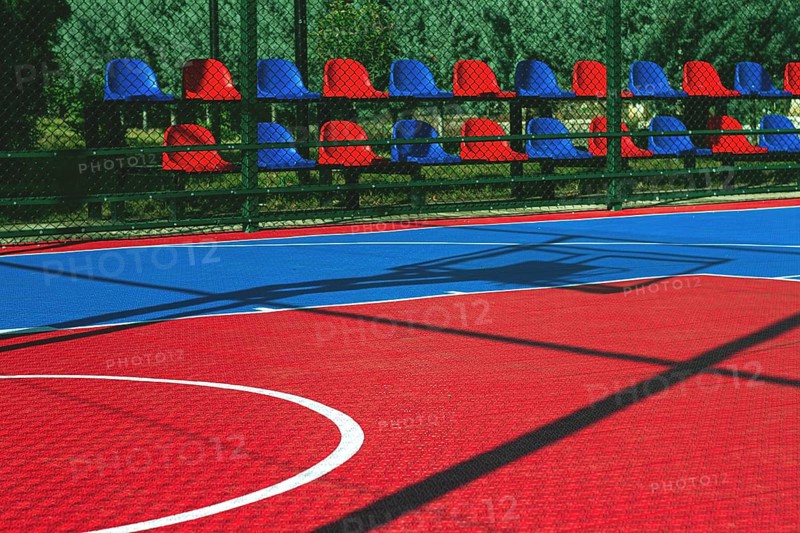 A vibrant basketball court features a mix of red and blue colors. Empty seats line the side, indicating a lively atmosphere earlier in the day. The sunlight casts interesting shadows on the surface