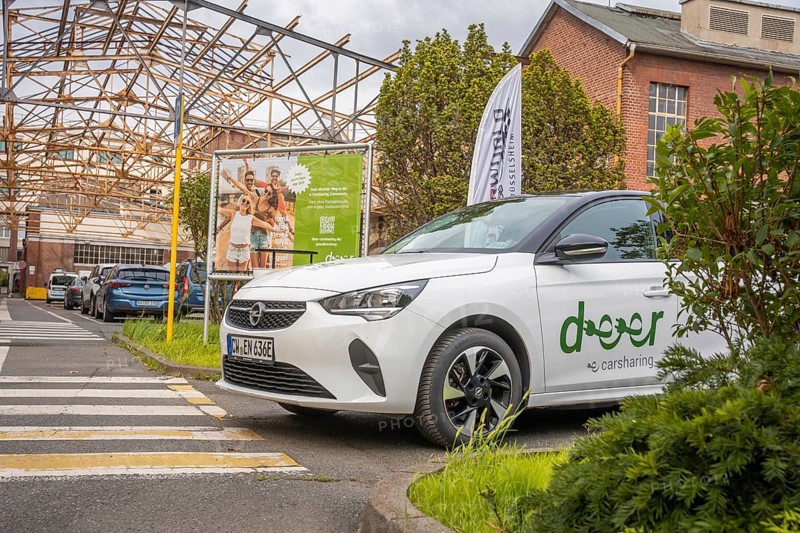 An Opel with car sharing logo stands in an urban environment with billboard and vegetation, Deer E-Car Sharing, Motorworld Rüsselsheim, Germany