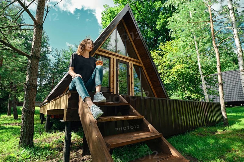 A woman sits on the steps of a wooden cabin surrounded by trees. She holds a drink and looks relaxed. The cabin has large windows and is in a sunny area. It is a calm day in the forest