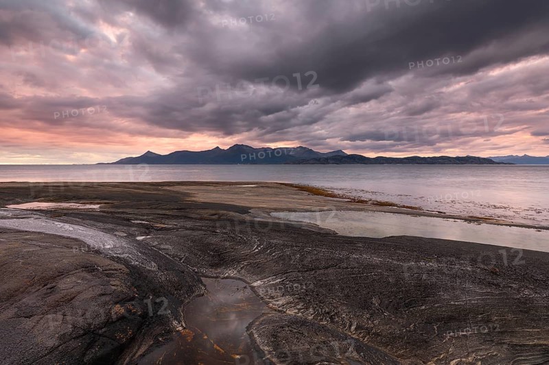 The Vestfjord in Nordland, Norway, offers a dramatic atmosphere near Bodø. Dark clouds are illuminated by the setting sun in shades of red and yellow. The rugged cliffs and mountains are reflected in the quiet fjord, which offers a breathtaking view of Lofoten