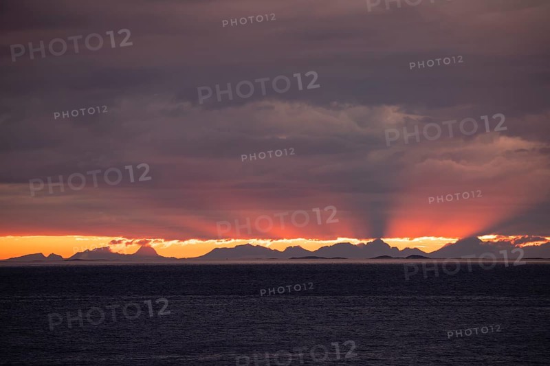 The Vestfjord in Nordland, Norway, offers a dramatic atmosphere near Bodø. Dark clouds are illuminated by the setting sun in shades of red and yellow. View towards Lofoten
