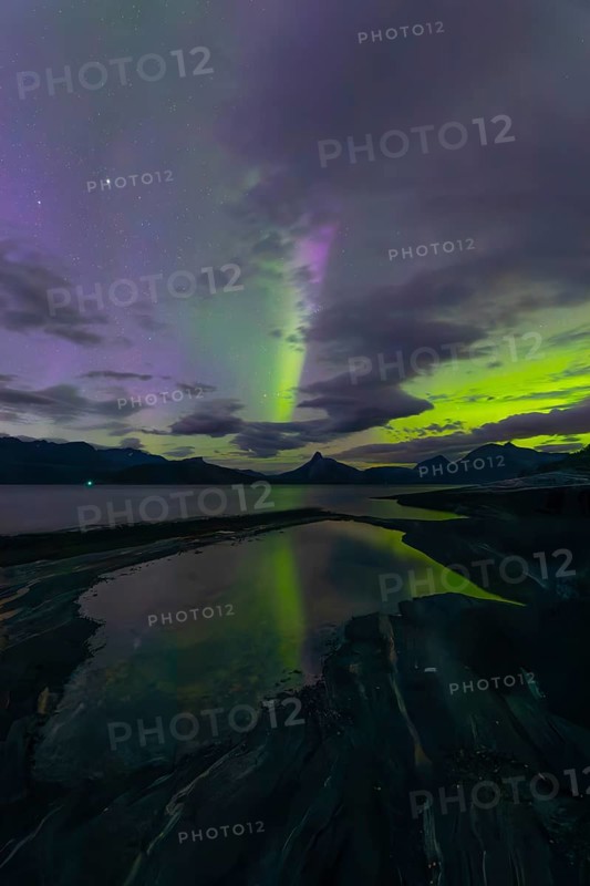 The Vestfjord in Nordland, Norway, has an enchanting atmosphere at night. Dark clouds and vivid aurora, polar lights dance over the fjord. The rugged cliffs and majestic mountains are reflected in the calm water and create a mystical backdrop