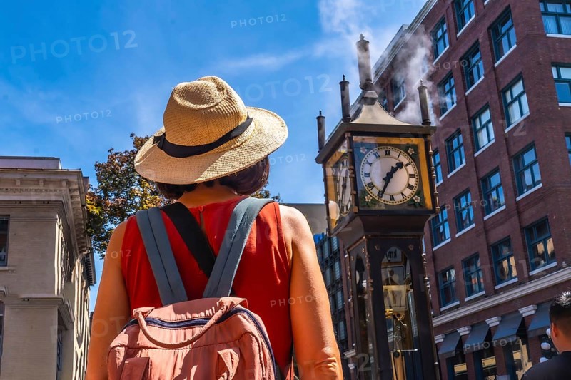 Female tourist wearing straw hat and backpack looking at the famous steam clock in gastown historic district, vancouver, british columbia, canada, in a sunny summer day