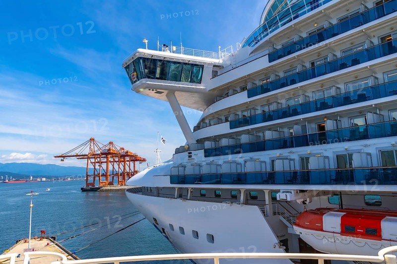 Large white cruise ship docking in vancouver harbor, surrounded by towering cargo cranes and majestic mountains under a bright blue sky on a sunny day, creating a picturesque scene