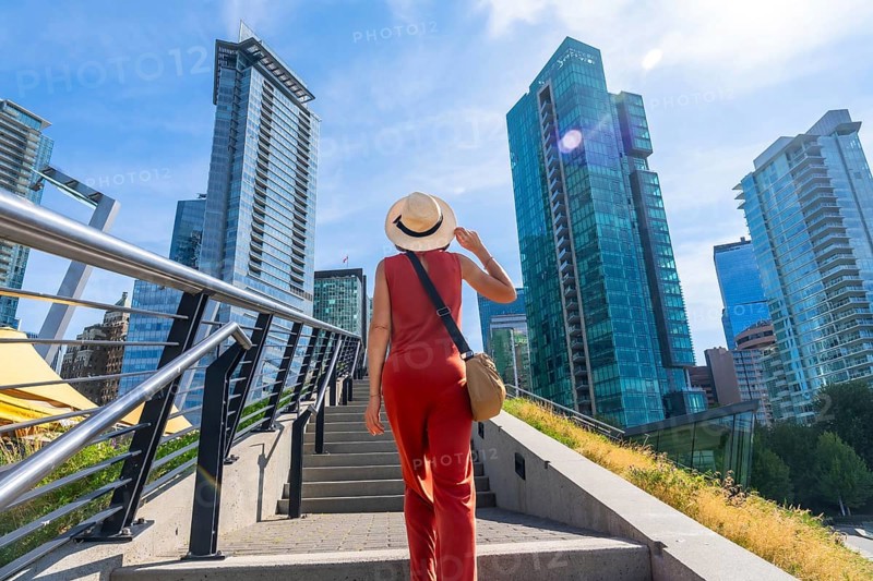 Female tourist wearing a straw hat and holding her hat is walking up stairs with modern glass skyscrapers in the background in vancouver, british columbia, canada