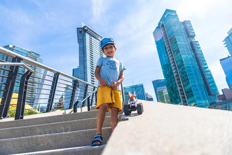 Young boy standing on steps playing with a toy car, with vancouver's modern buildings and clear blue sky as a backdrop, enjoying a sunny day in the city