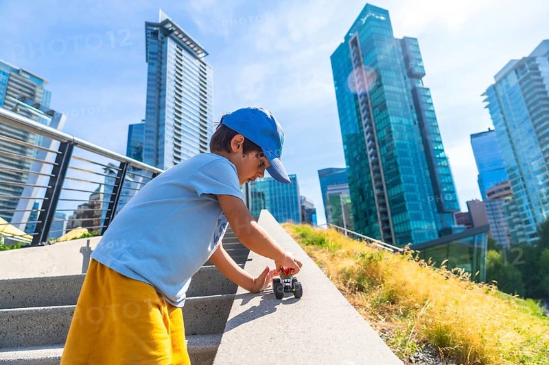 Young boy playing joyfully with a toy car on the steps of an urban park, soaking up the warmth of a sunny day, with vancouver's impressive skyline rising in the background