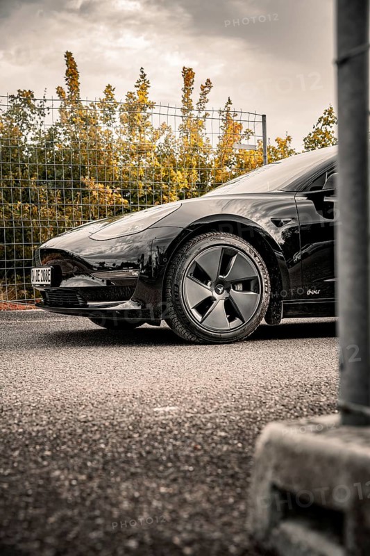 Black car on asphalt in front of a fence surrounded by trees and clouds, Deer E-Carsharing, Tesla Model 3, Calw, Germany