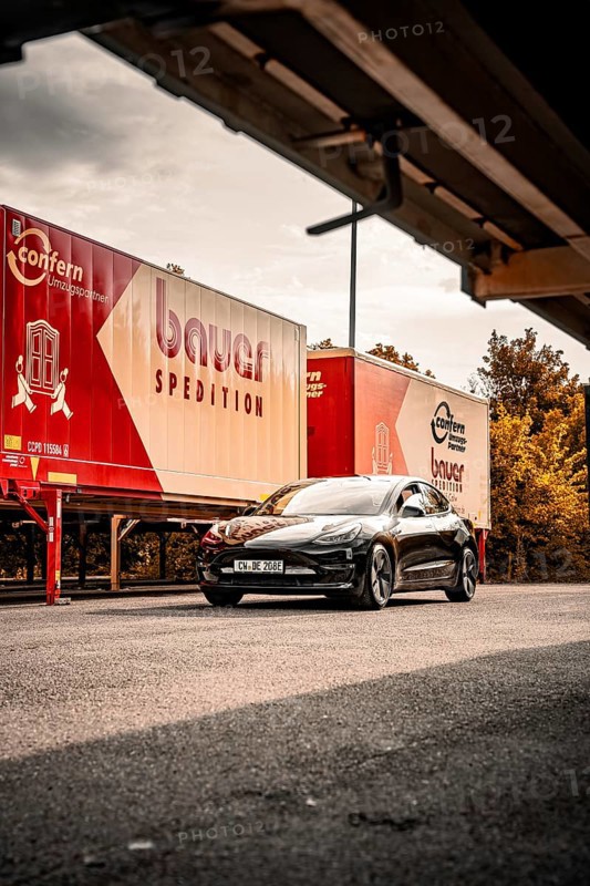Black car in front of trucking trailers on a road with cloudy sky and trees, Deer E-Carsharing, Tesla Model 3, Calw, Germany