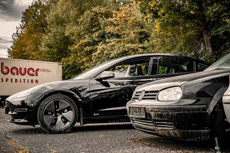 Black car and older vehicle in a parking lot with a truck in the background, Deer E-Carsharing, Tesla Model 3, Calw, Germany