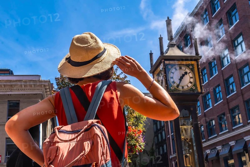 Young woman with backpack and straw hat looking at the famous steam clock in gastown, vancouver, enjoying a sunny summer day exploring the historic district of british columbia