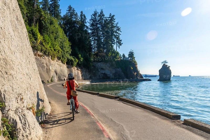 Tourist is enjoying a sunny day riding a bicycle along the stanley park seawall, with the iconic siwash rock and cargo ships in the background, in vancouver, british columbia
