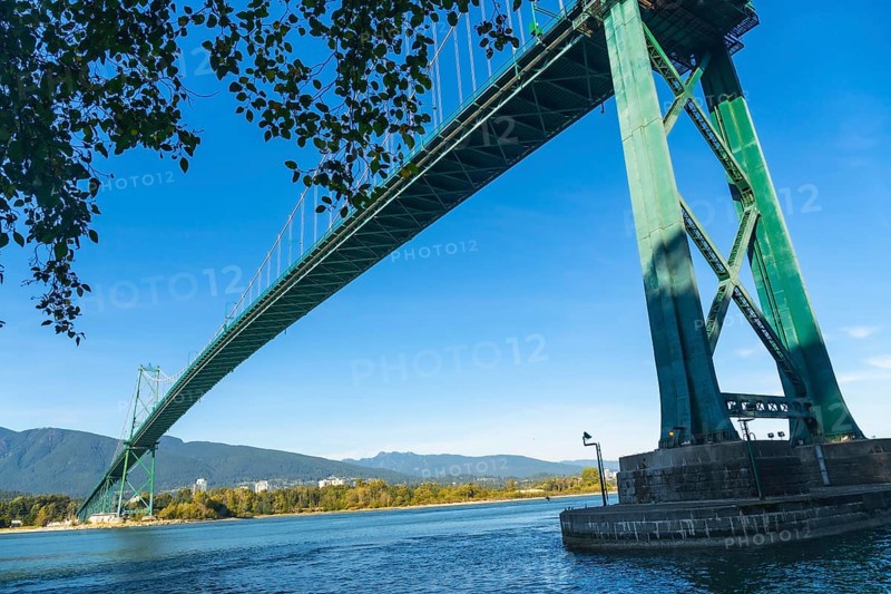 Lions gate bridge extending over burrard inlet with stanley park seawall in foreground and north shore mountains in background on clear sunny day in vancouver, british columbia