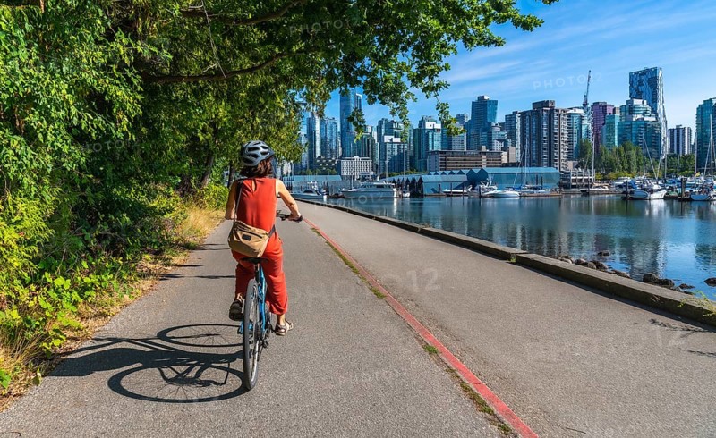 Tourist wearing helmet and orange jumpsuit is riding bicycle on seawall in stanley park with vancouver city skyline, marina and sea in background on sunny summer day
