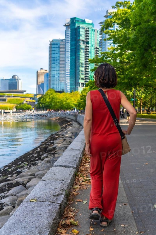 Female tourist wearing red jumpsuit walking along waterfront promenade in vancouver, british columbia, enjoying cityscape view on sunny summer day