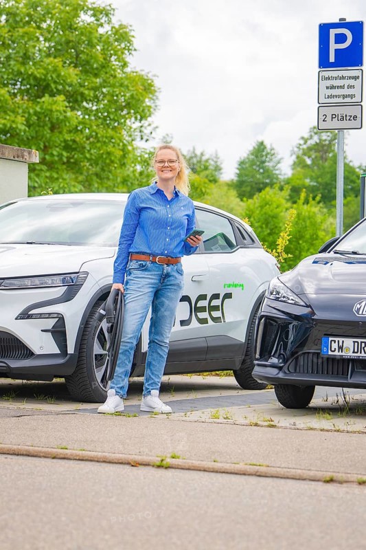 Woman standing in front of two electric cars in a parking lot with technological flair, Deer E-Carsharing, Calw, Germany