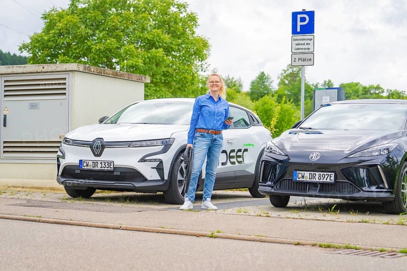 Two electric cars park side by side at a charging station, surrounded by green countryside, Deer E-Carsharing, Calw, Germany