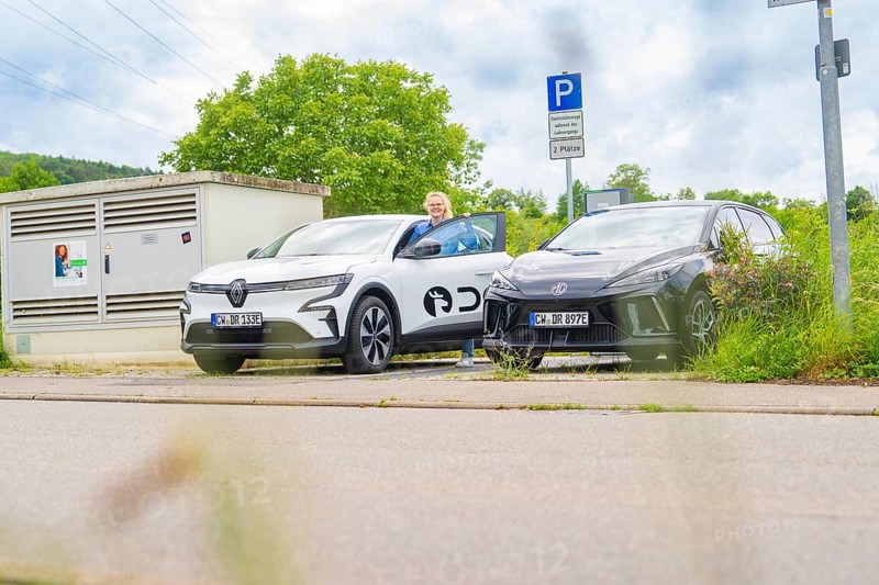 Two electric cars are parked in a parking lot, and a man is standing next to them. Cloudy sky and green surroundings, Deer E- Carsharing, Calw, Germany