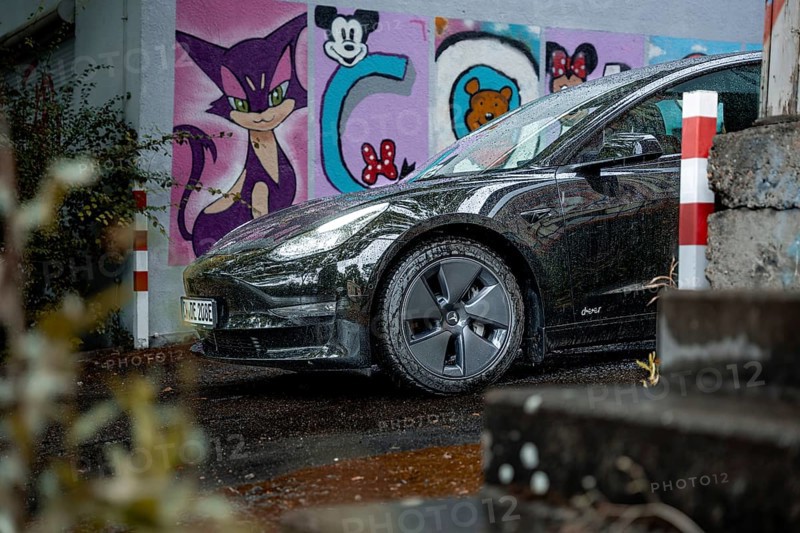 Black car in an urban environment with colorful graffiti wall and wet road, Deer e-Carsharing, Tesla Model 3, Calw, Germany