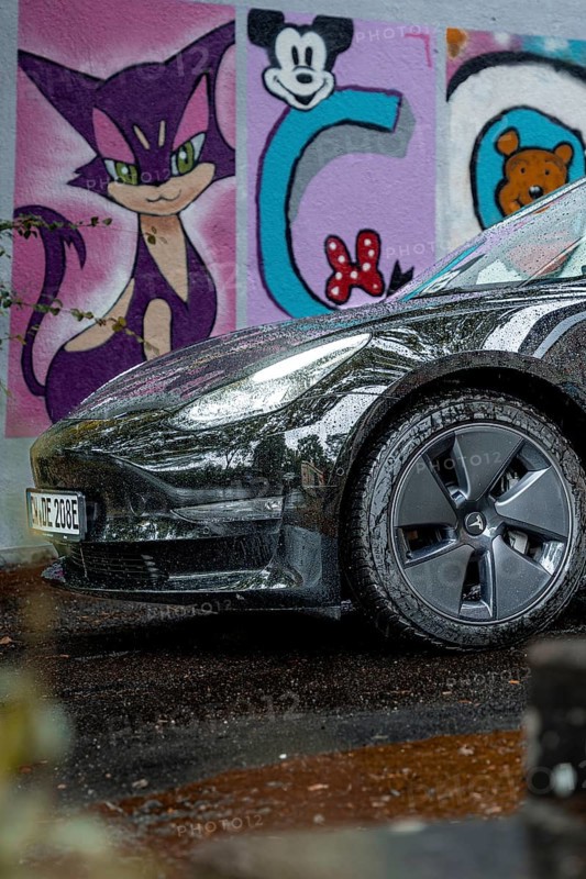 Close-up view of a black car in front of colorful graffiti art on a wet road, Deer e-car sharing, Tesla Model 3, Calw, Germany