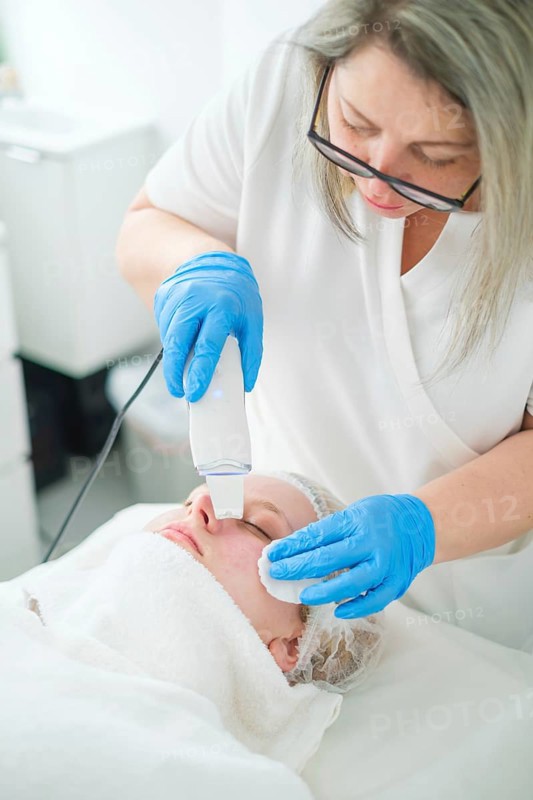 A pro esthetician cosmetologist uses a metal spatula for ultrasonic facial cleansing to exfoliate and refresh the client's skin