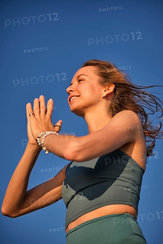A woman practices yoga on the beach during sunset, holding her hands in Namaskar Mudra while smiling