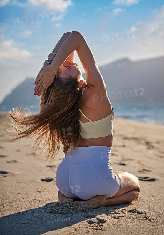 Woman performs Vajrasana with backbend on sandy beach as the sun sets over the ocean. She kneels on the sand with arms raised