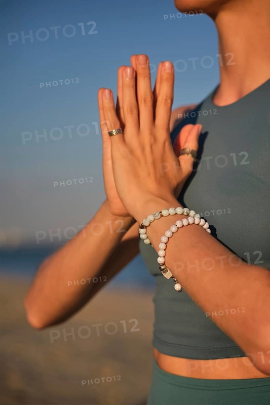 A woman practices yoga by holding hands in Namaskar Mudra at the beach as the sun sets in the background, creating a stunning scene