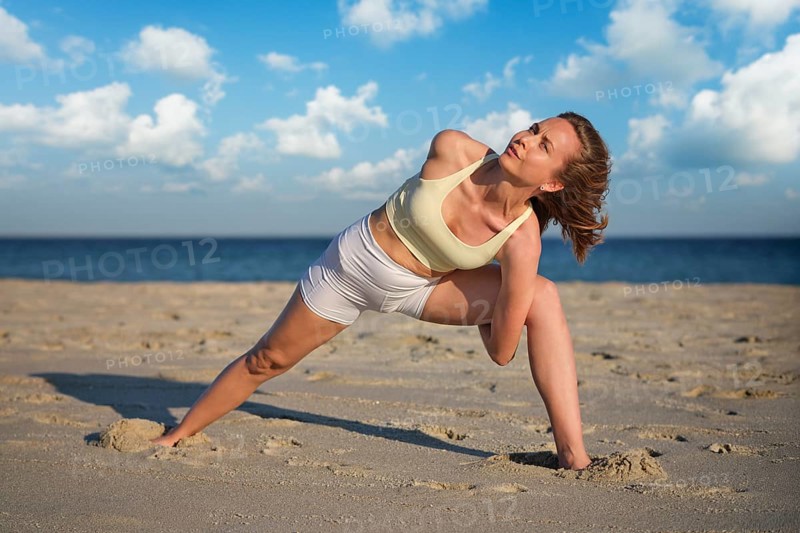 A woman practices Baddha Trikonasana on a sandy beach with wide legs and arms bound. The sun sets in the background