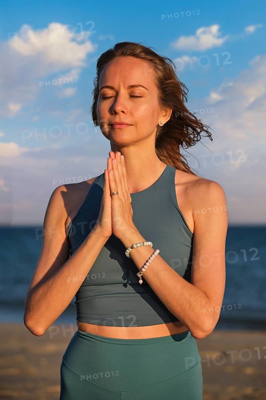 A woman practices yoga on the beach at sunset, holding hands in Namaskar Mudra as she meditates with her eyes closed