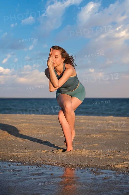 A woman practices standing Garudasana on a beach with one leg wrapped around the other at sunset by the water