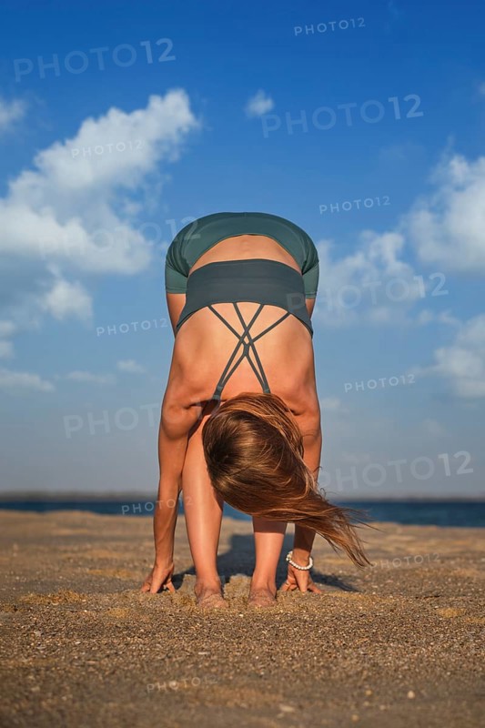 A woman does Uttanasana on a sandy beach during sunset, focusing on her breath and stretching her body forward