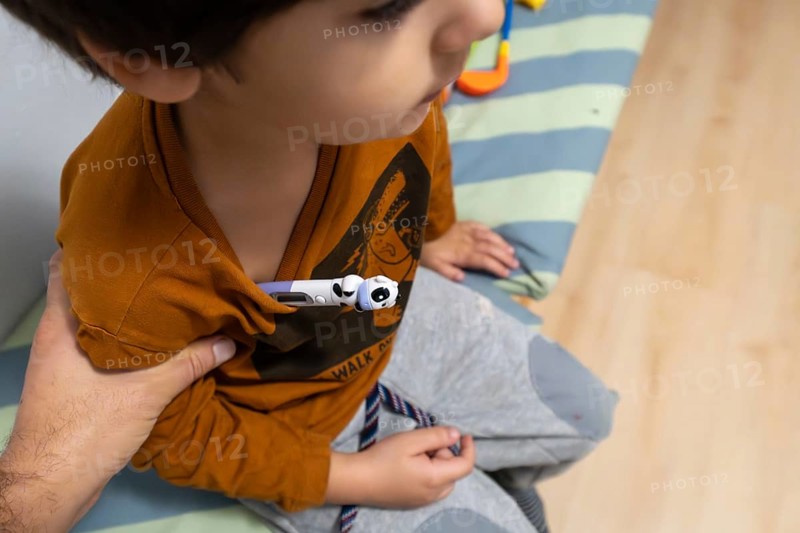 Child wearing a t shirt having a digital thermometer placed under arm for temperature measurement, indicating illness, flu, or a fever, with a parents arm providing assistance and comfort