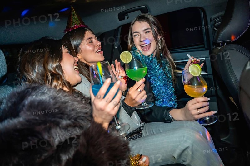 Young happy women friends are enjoying a new year's eve celebration or party inside a car, toasting with colorful cocktails, laughing, and having fun together at night