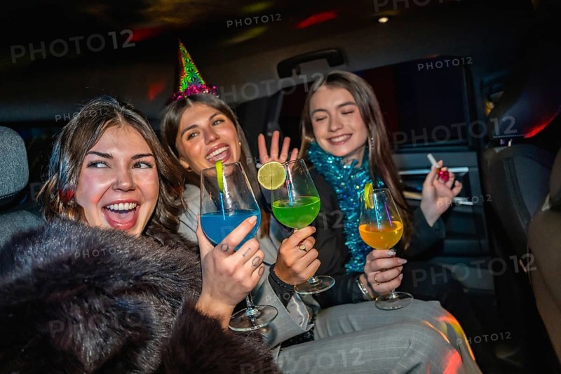 Young women friends celebrating new year's eve, cheering with colorful cocktails while traveling in the back seat of a car at night, enjoying the festive atmosphere