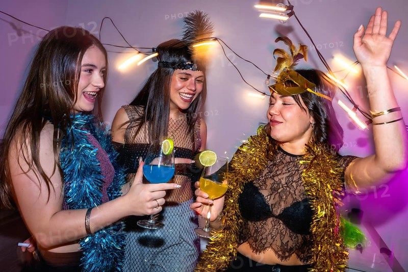 Three young women smiling, laughing, and drinking colorful cocktails while having fun at a festive new year's eve or costume party, surrounded by sparkling string lights and tinsel decorations