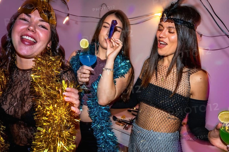 Young women in vibrant costumes laugh and clink cocktails at a festive new year's eve party indoors, celebrating friendship, glamour, and joyful togetherness late at night