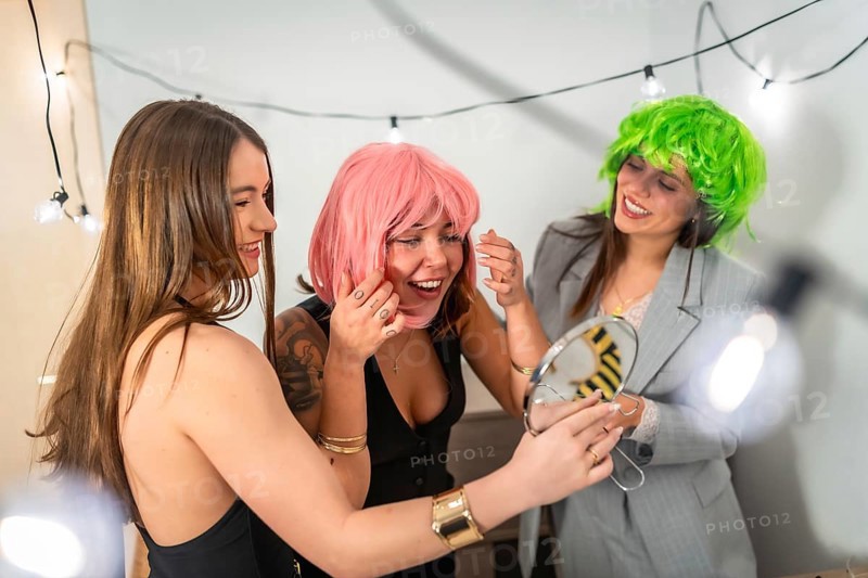 Group of happy young women friends trying on fun colorful wigs and preparing for a costume party or new year's eve celebration at home, sharing moments of joy and friendship
