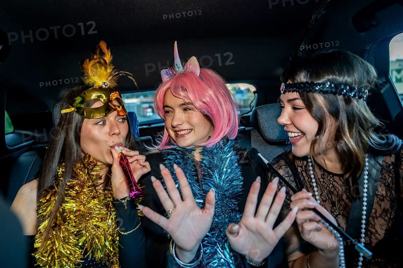 Three young women in festive costumes ride in a car, wearing tinsel and makeup, laughing and blowing party blowers as they celebrate a joyful night out together