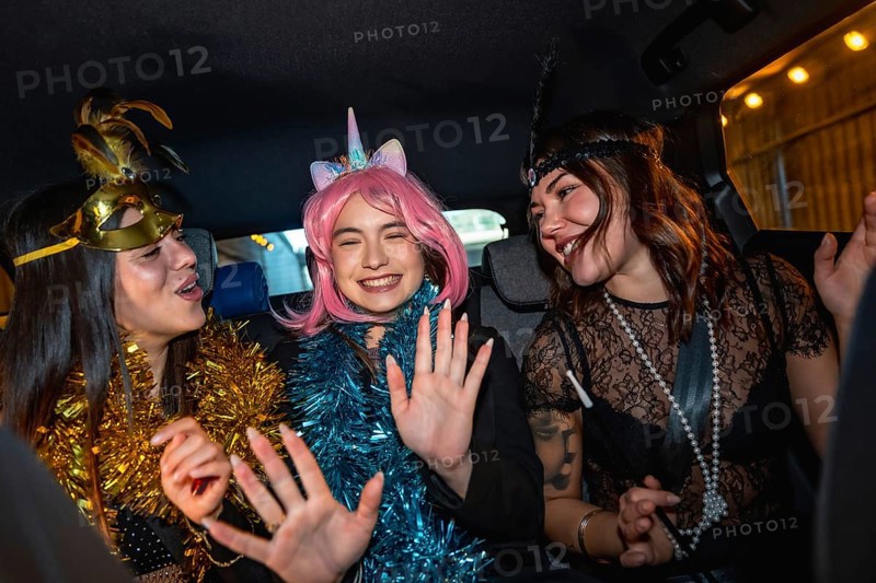 Three young women wearing festive costumes and garlands are happily laughing and dancing while safely traveling in the back seat of a car during a night celebration