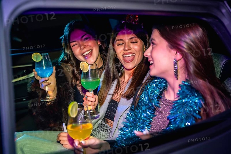 Three friends joyfully celebrating a festive occasion, laughing and holding colorful cocktails inside a car, enjoying a night out together with vibrant energy
