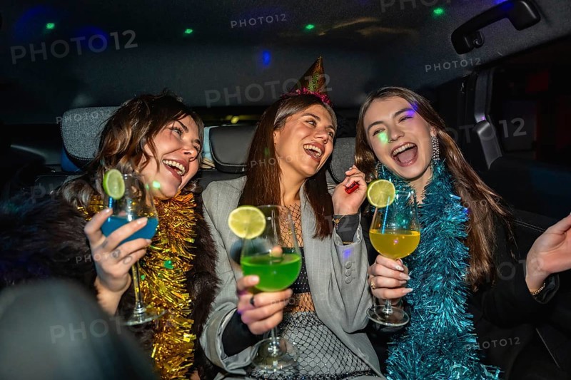 Three friends dressed up in party hats and tinsel garlands laughing and toasting colorful cocktails inside a car, enjoying a festive night out full of joy and spontaneous fun