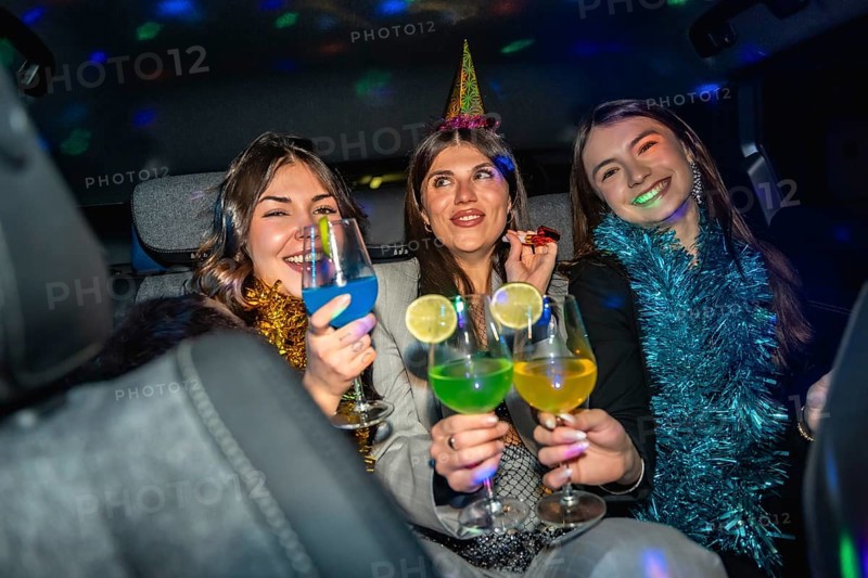 Three smiling young women wearing festive hats and tinsel, holding colorful cocktails, enjoying a night out while disco lights illuminate the car interior