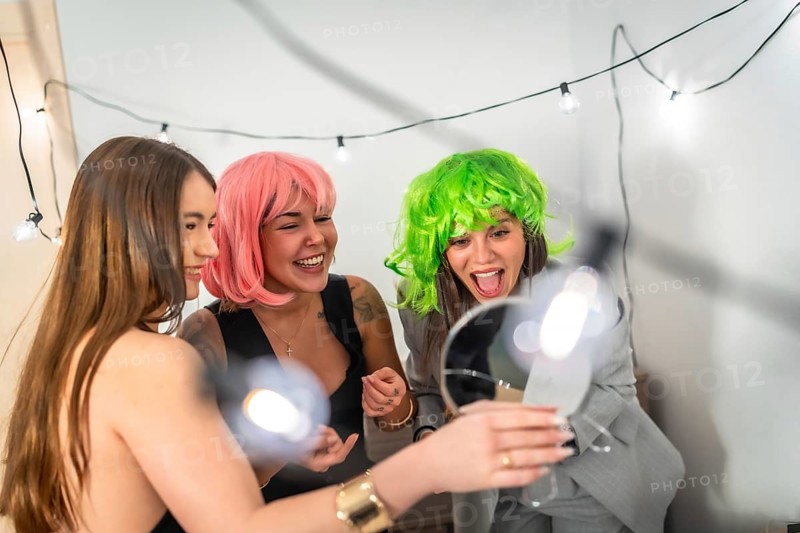 Two young women in pink and green wigs laugh and celebrate at a colorful house party, sharing joyful candid moments, friendship and playful festive fun indoors with lights and decorations