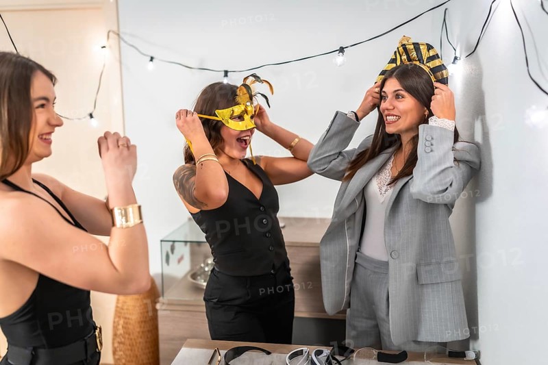 Happy young women enjoying a costume party at home, laughing and having fun while trying on masks and festive headbands, celebrating holidays together