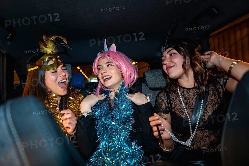 Three young women smiling, laughing, and having fun, dressed up in costumes with festive accessories, enjoying a lively party ride in the back seat of a car at night