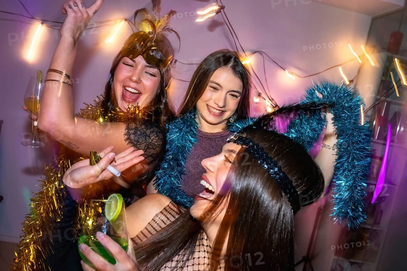 Three young women laughing and dancing, celebrating a festive new year's eve or costume party at home, wearing fun accessories and drinking cocktails in a lively atmosphere