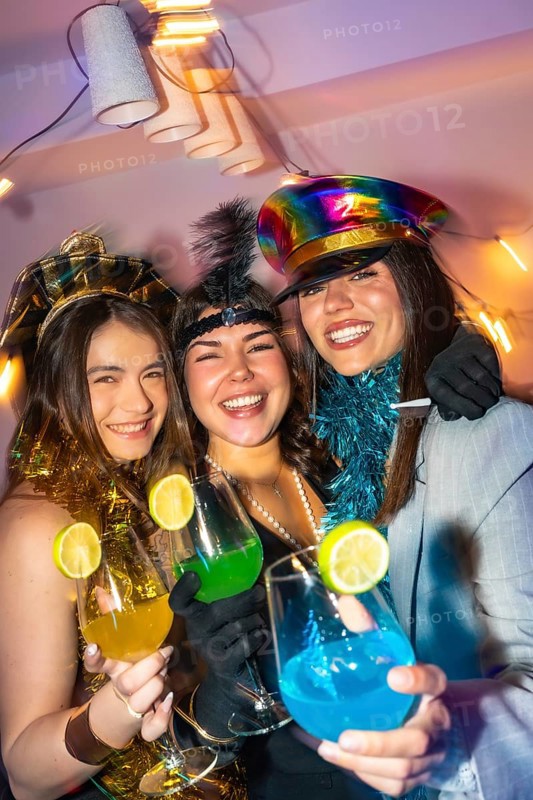 Three happy young women celebrating new year's eve, wearing festive costumes and holding colorful drinks with lime garnishes in a lively, vibrant party atmosphere